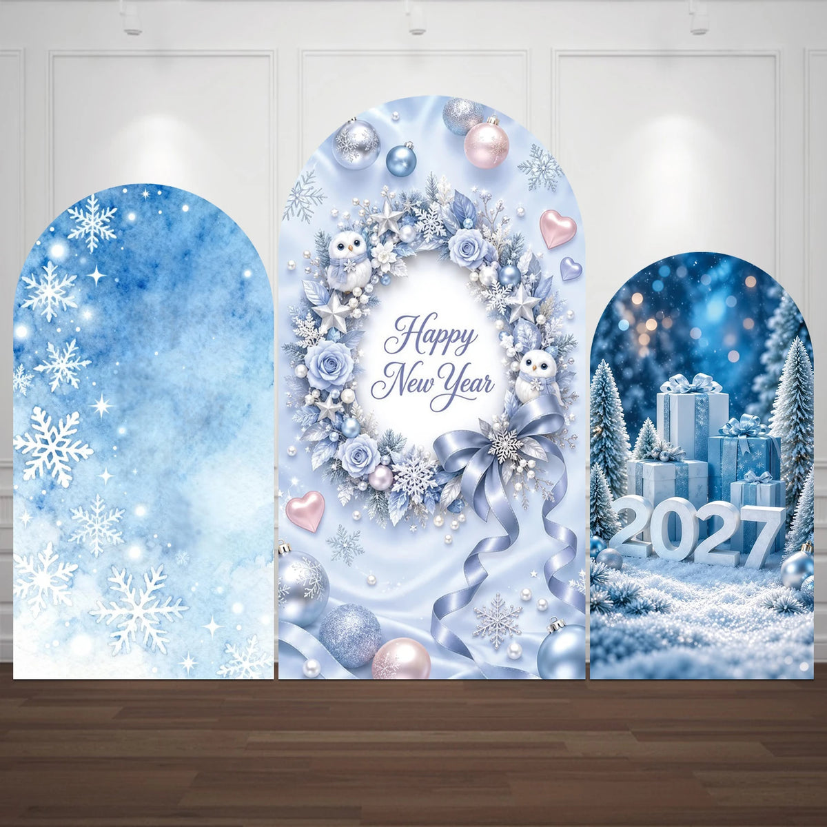 Three arched blue New Year backdrop boards with winter decorations, displayed on a wooden floor against a white paneled wall. The left board has snowflakes and a blue gradient, the center says 'Happy New Year' with a floral wreath and snow owl, and the right shows snow, gifts, trees, and '2027'.
